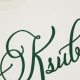 Close-up of the KSUBI Prestige Biggie Short Sleeve T-shirt in antique white, featuring large dark green cursive embroidery across the front. The premium cotton jersey collar and part of the KSUBI neck label are also visible.