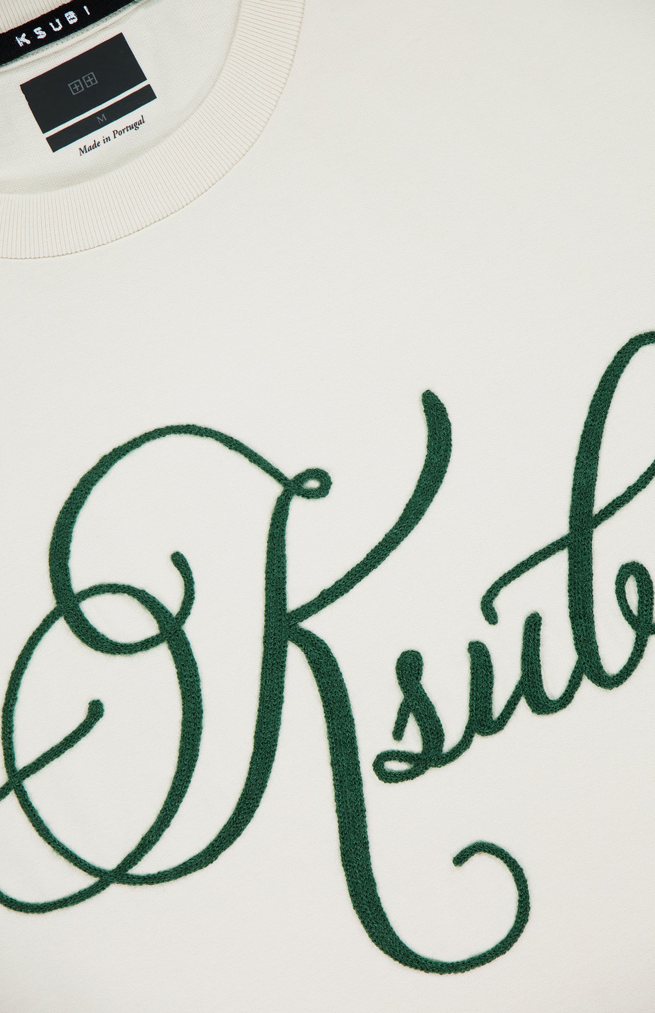 Close-up of the KSUBI Prestige Biggie Short Sleeve T-shirt in antique white, featuring large dark green cursive embroidery across the front. The premium cotton jersey collar and part of the KSUBI neck label are also visible.