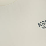 Close-up of the beige KSUBI State Halen Cut Off T-shirt featuring a ribbed collar and small gray KSUBI EST logo on the chest. The neck label and part of the inside tag are also visible.