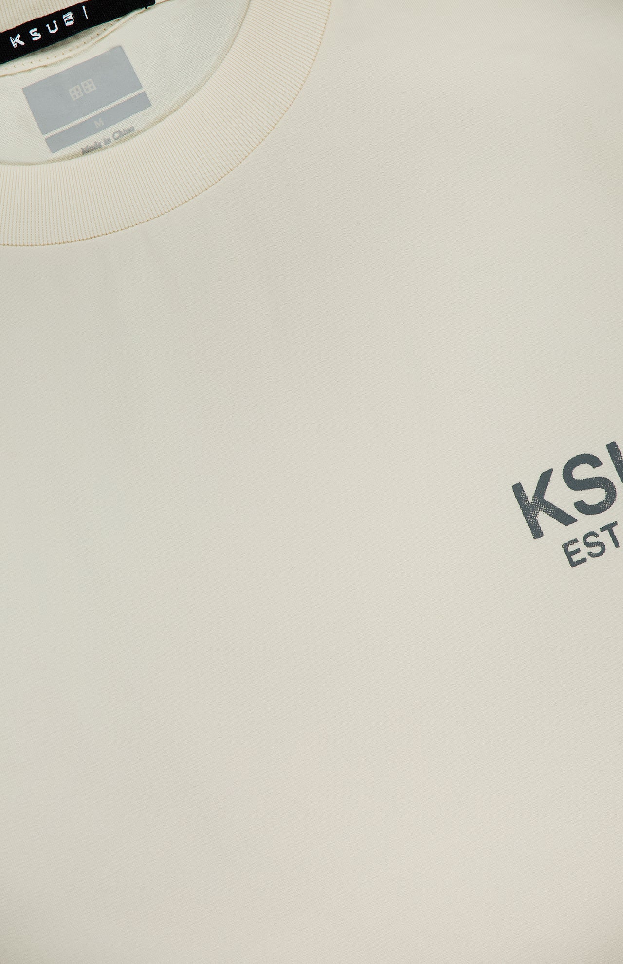 Close-up of the beige KSUBI State Halen Cut Off T-shirt featuring a ribbed collar and small gray KSUBI EST logo on the chest. The neck label and part of the inside tag are also visible.