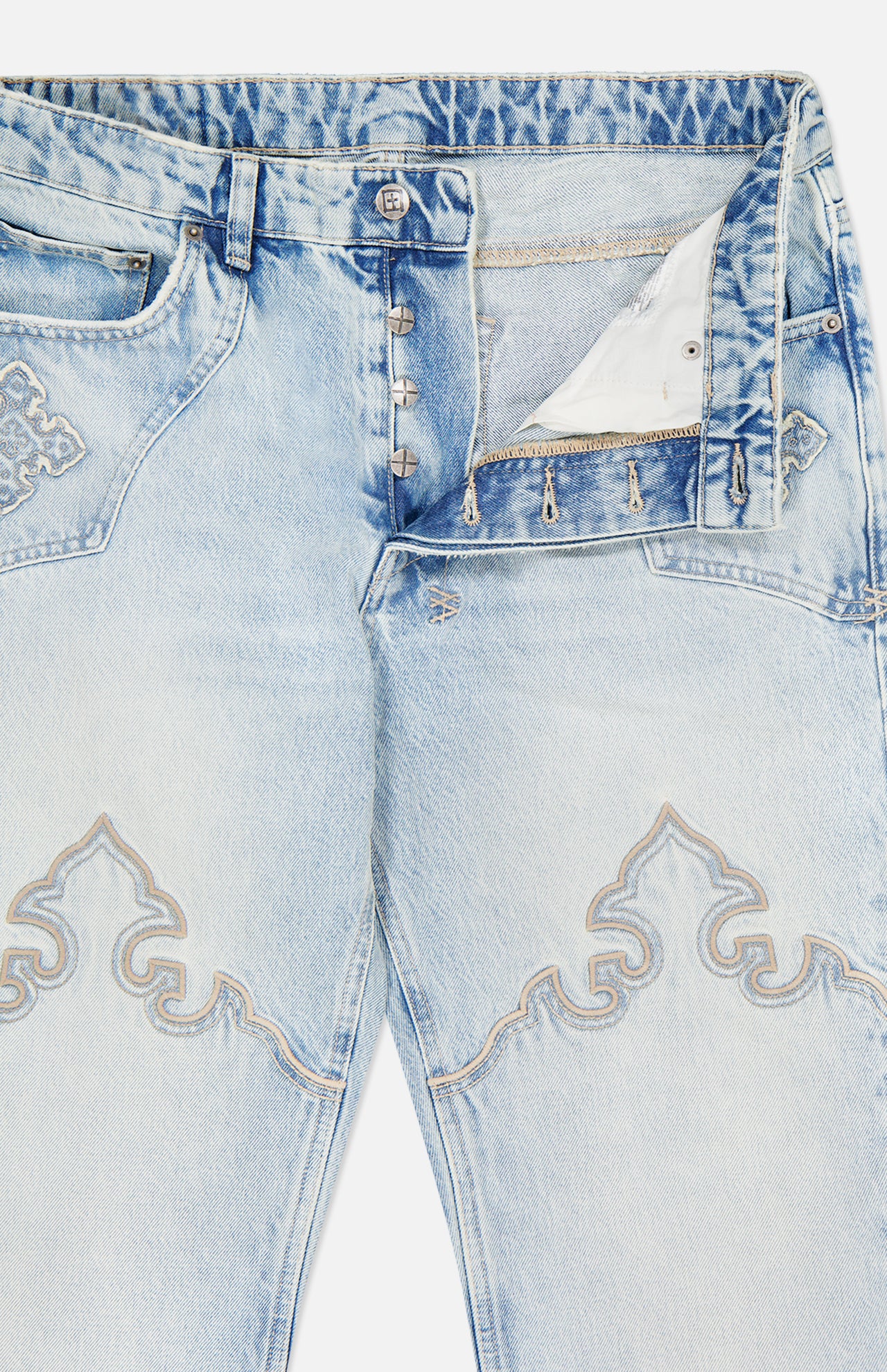 KSUBI Toro Western Knee Paneling Jeans in light blue feature a silver-toned button fly, intricate stitched patterns on the thighs and pocket area, and a slightly faded, distressed finish. The partially open fly reveals white pocket lining, while classic embroidered denim details lend a subtle western flair.