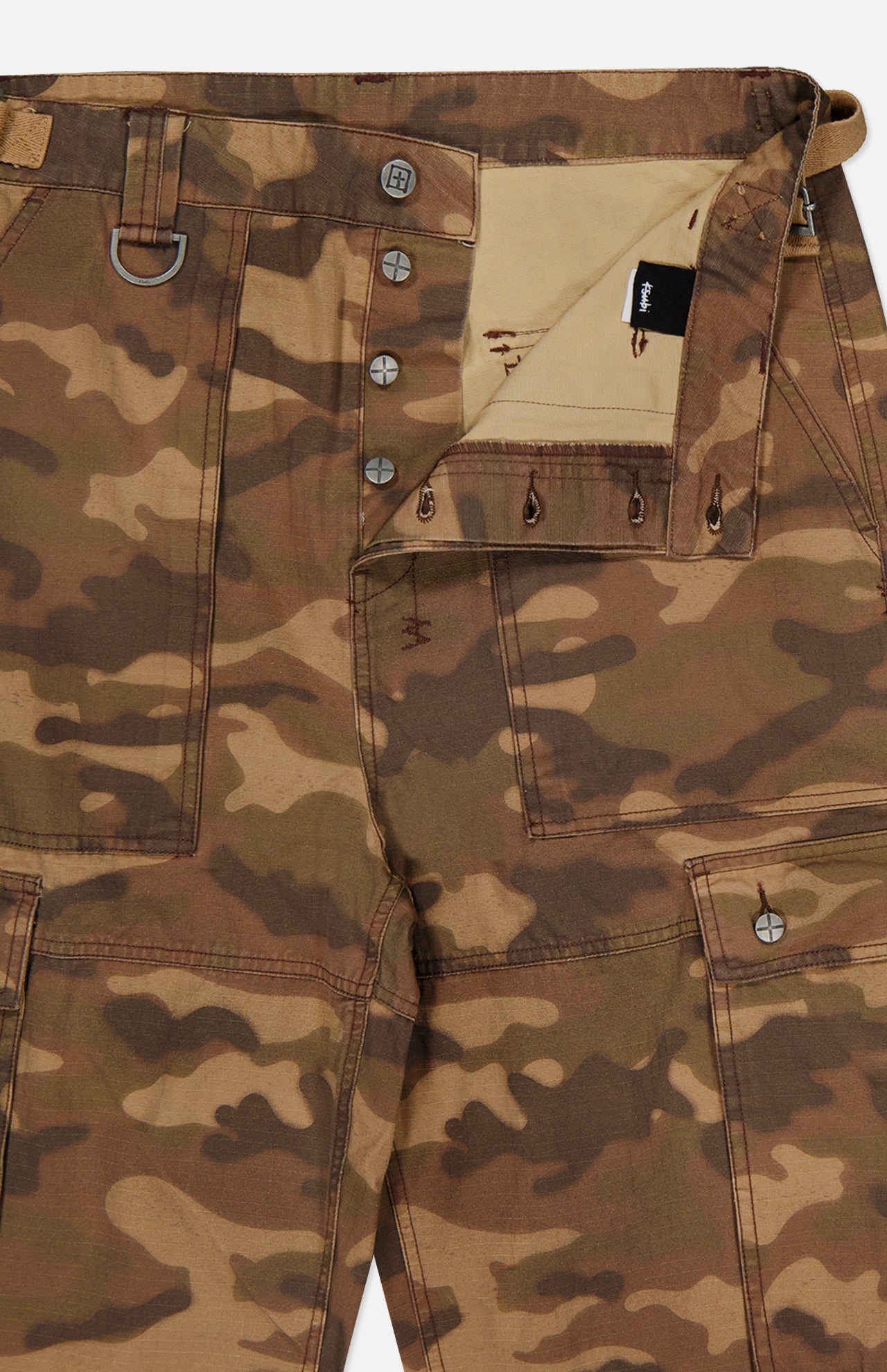 Close-up of the KSUBI Troop Camouflage Cargo Pant, crafted from durable cotton twill in brown and olive camo, featuring large front pockets, a partially open button fly, a visible waistband, and a metal D-ring detail on the left side.