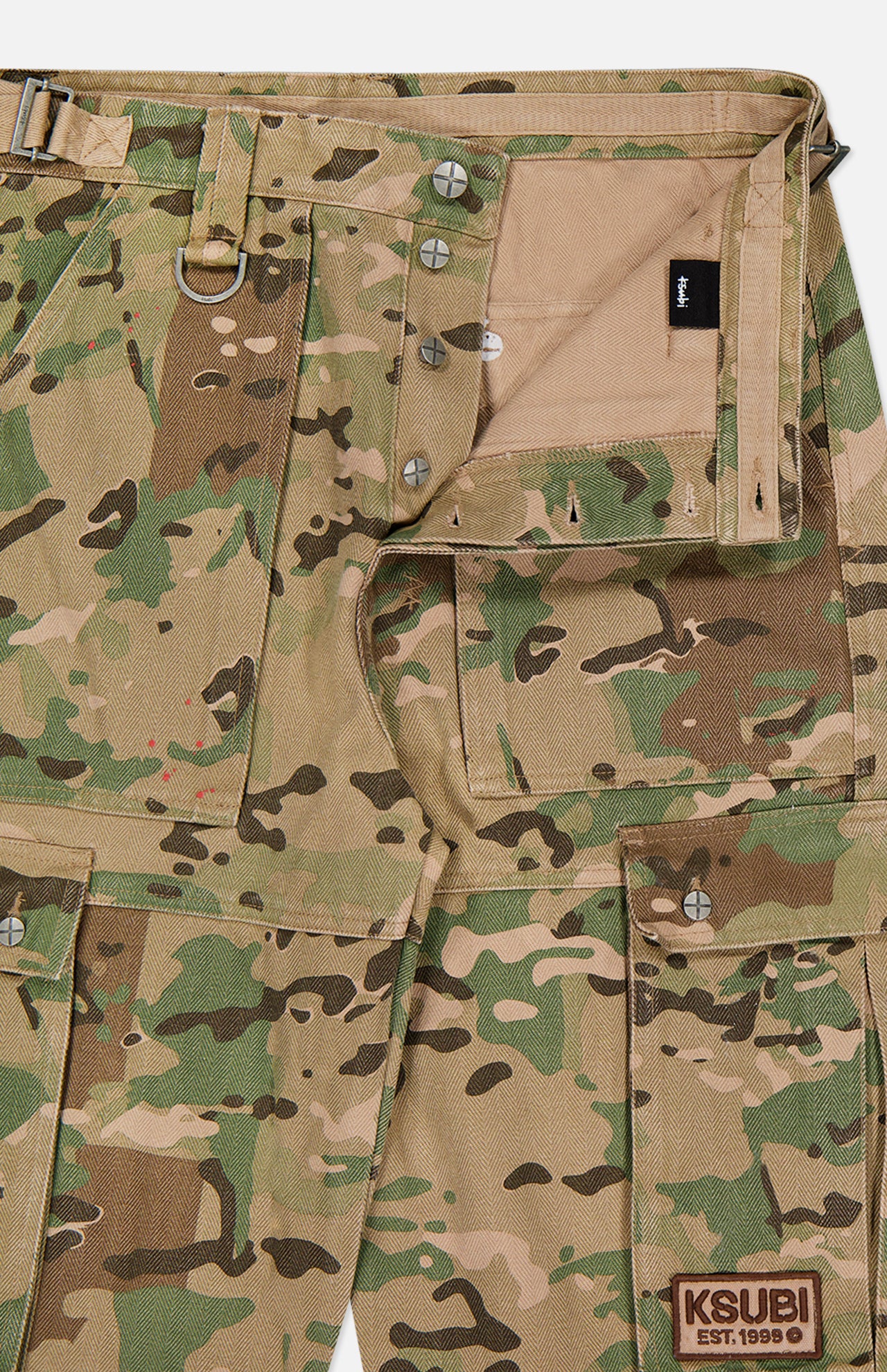 Close-up of the KSUBI Trooper Camouflage Cargo Pant featuring woodland camo, metal buttons, belt loops, multiple pockets, and a visible KSUBI patch above the left knee. The open fly reveals the inside label and relaxed leg fit.
