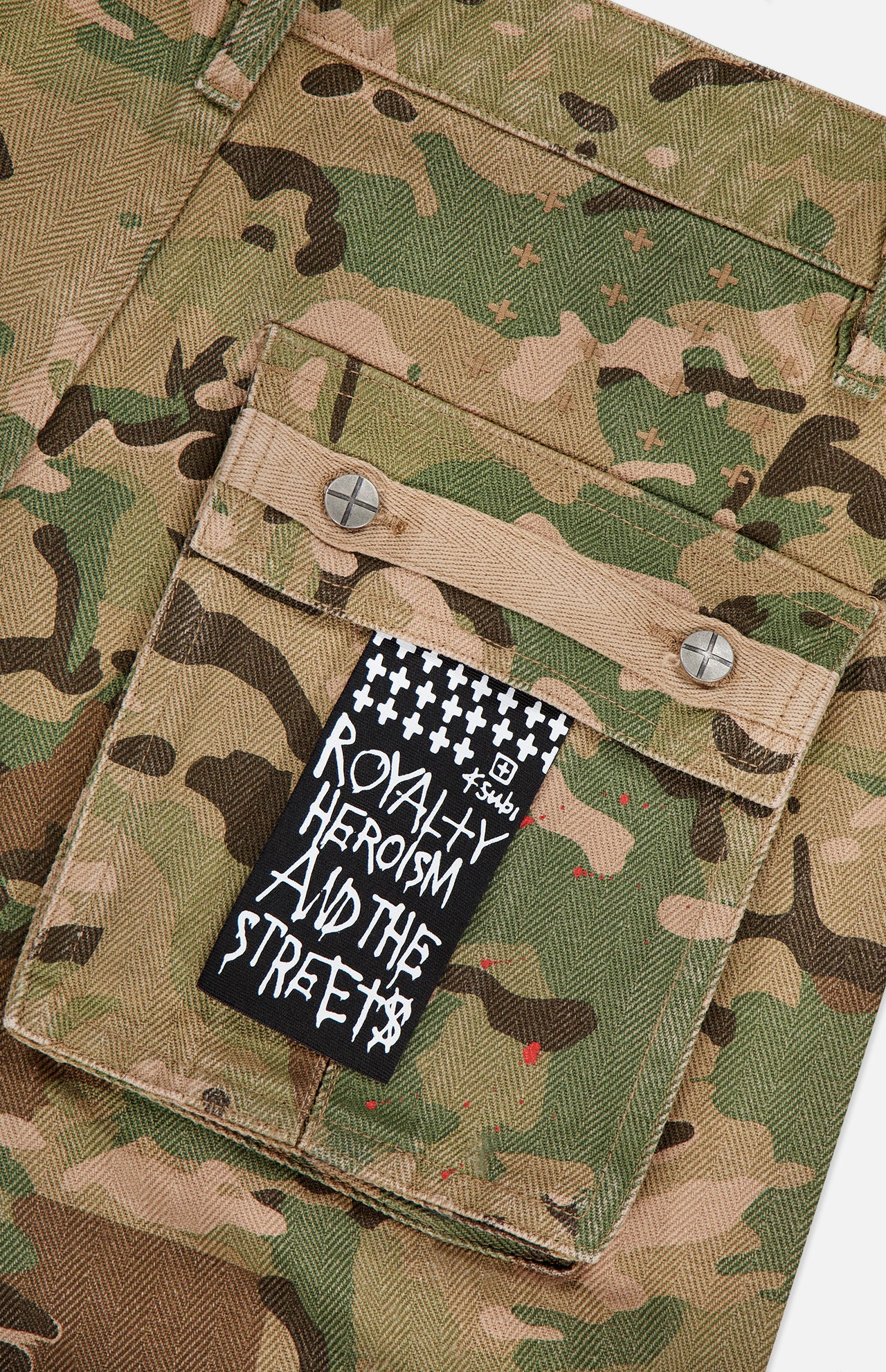 A close-up of the KSUBI Trooper Camouflage Cargo Pant pocket, featuring two metal rivets and a black and white label with "ROYALTY HEROISM AND THE STREETS" in bold, graffiti-style text.