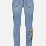 KSUBI Van Winkle Duke 4Eva Jeans in light blue comfort stretch denim, shown from the back, featuring two back pockets, a small brown patch, distressed detailing near the waistband, and a bold yellow rose with green leaves embroidered on one leg.