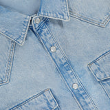 The KSUBI Whiplash Long Sleeve Western Shirt is crafted from light blue denim and features silver snap buttons, a pointed collar, and two front chest pockets with flaps. This shirt showcases a subtle faded texture throughout the fabric, while the top of a black KSUBI label is partly visible at the neckline.