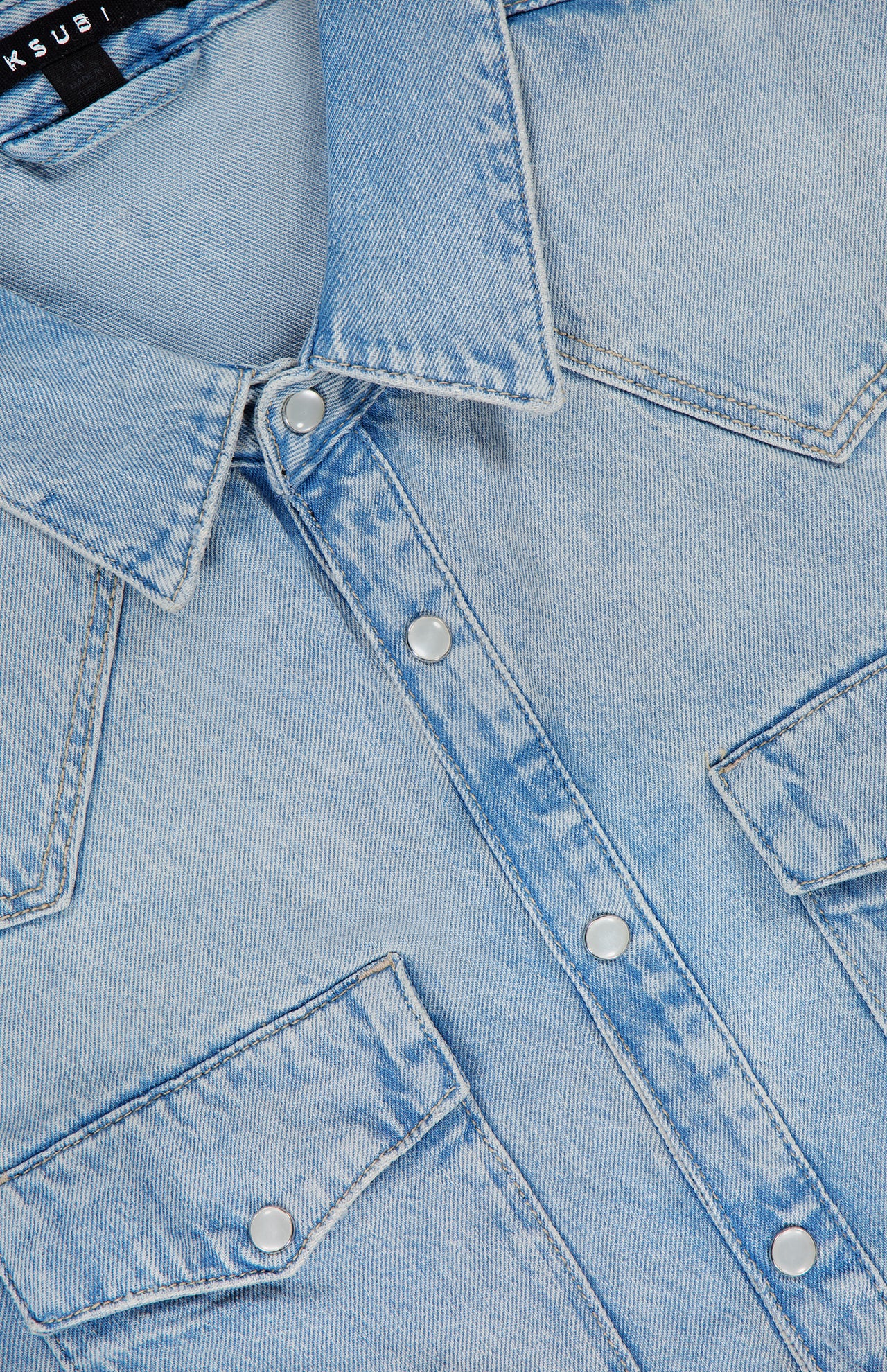 The KSUBI Whiplash Long Sleeve Western Shirt is crafted from light blue denim and features silver snap buttons, a pointed collar, and two front chest pockets with flaps. This shirt showcases a subtle faded texture throughout the fabric, while the top of a black KSUBI label is partly visible at the neckline.