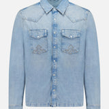 The Whiplash Long Sleeve Western Shirt by KSUBI features a light blue, slightly faded denim fabric, long sleeves, a pointed collar, button-down front, and two chest pockets with flap closures. Its casual finish gives this classic western shirt a relaxed yet timeless appeal.