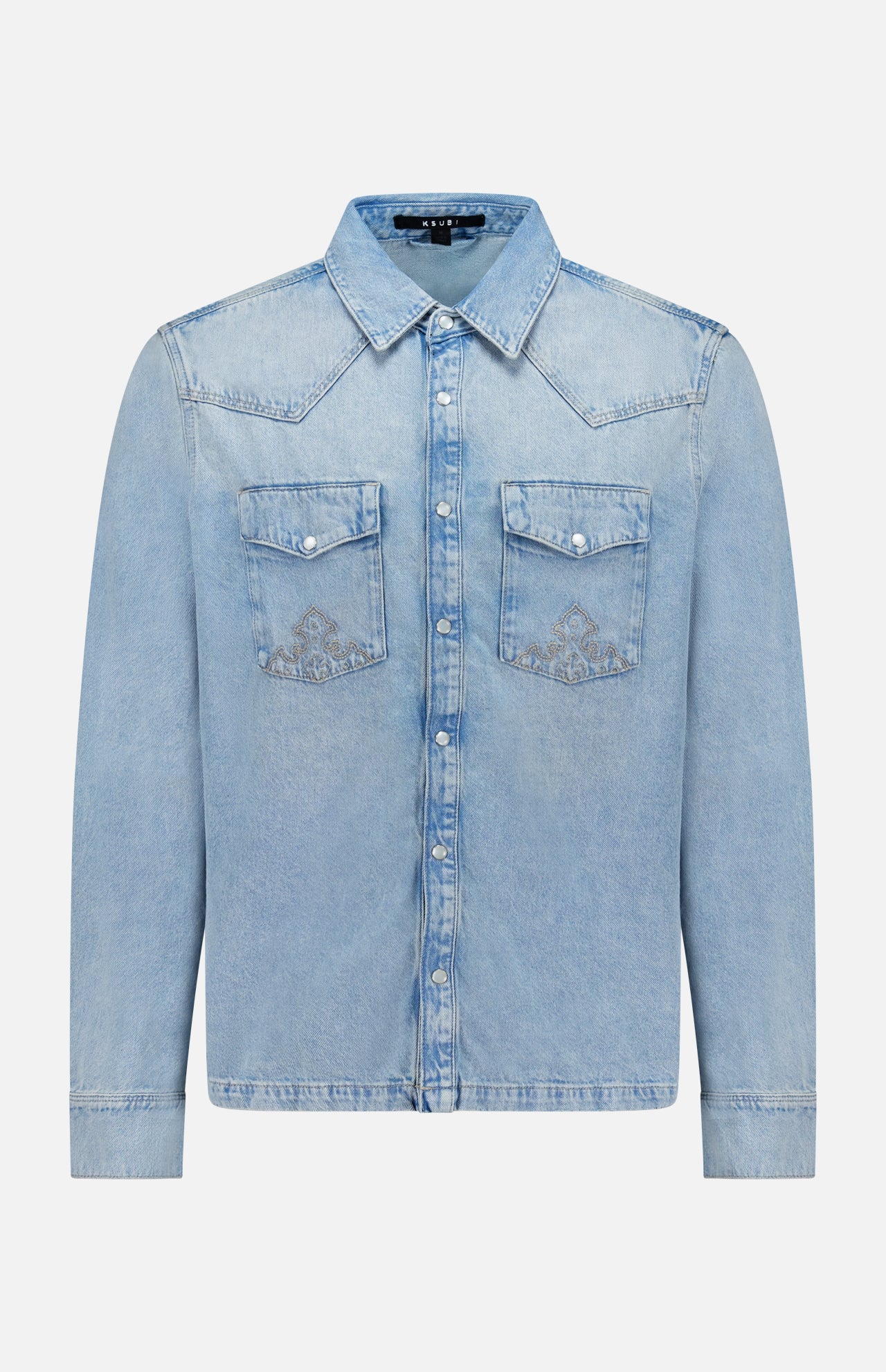 The Whiplash Long Sleeve Western Shirt by KSUBI features a light blue, slightly faded denim fabric, long sleeves, a pointed collar, button-down front, and two chest pockets with flap closures. Its casual finish gives this classic western shirt a relaxed yet timeless appeal.