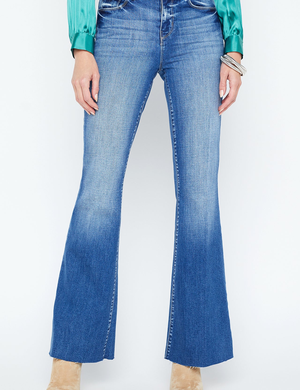 A person wearing L'AGENCE Sera High Rise Sneaker Flare Jeans, a teal satin long-sleeve blouse, and beige pointed-toe heeled boots stands against a white background.
