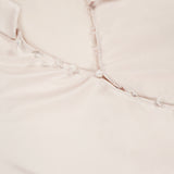 Close-up of the Bernice Satin Wrap Blouse by L’AGENCE in pale pink champagne, featuring a collar, delicate fabric-covered buttons down the front, and a visible L'AGENCE brand label on the inside neckline.