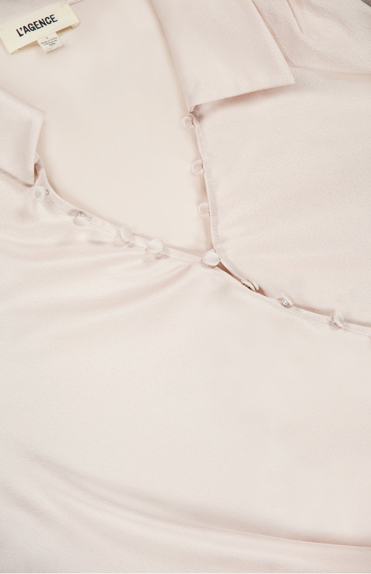 Close-up of the Bernice Satin Wrap Blouse by L’AGENCE in pale pink champagne, featuring a collar, delicate fabric-covered buttons down the front, and a visible L'AGENCE brand label on the inside neckline.