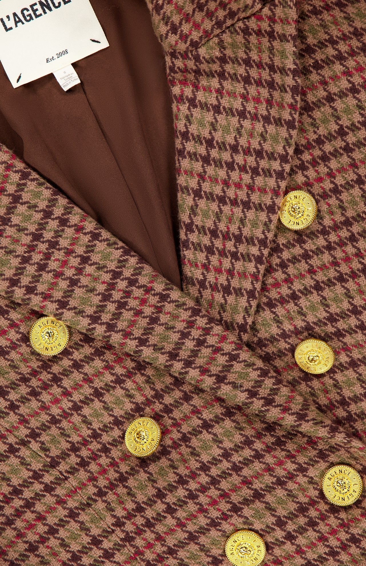 Close-up of the L’AGENCE Bethany Structured Houndstooth Blazer in brown and burgundy plaid, featuring structured shoulders, gold decorative buttons, brown lining, and an inner label that reads “L’AGENCE Est. 2008.”.