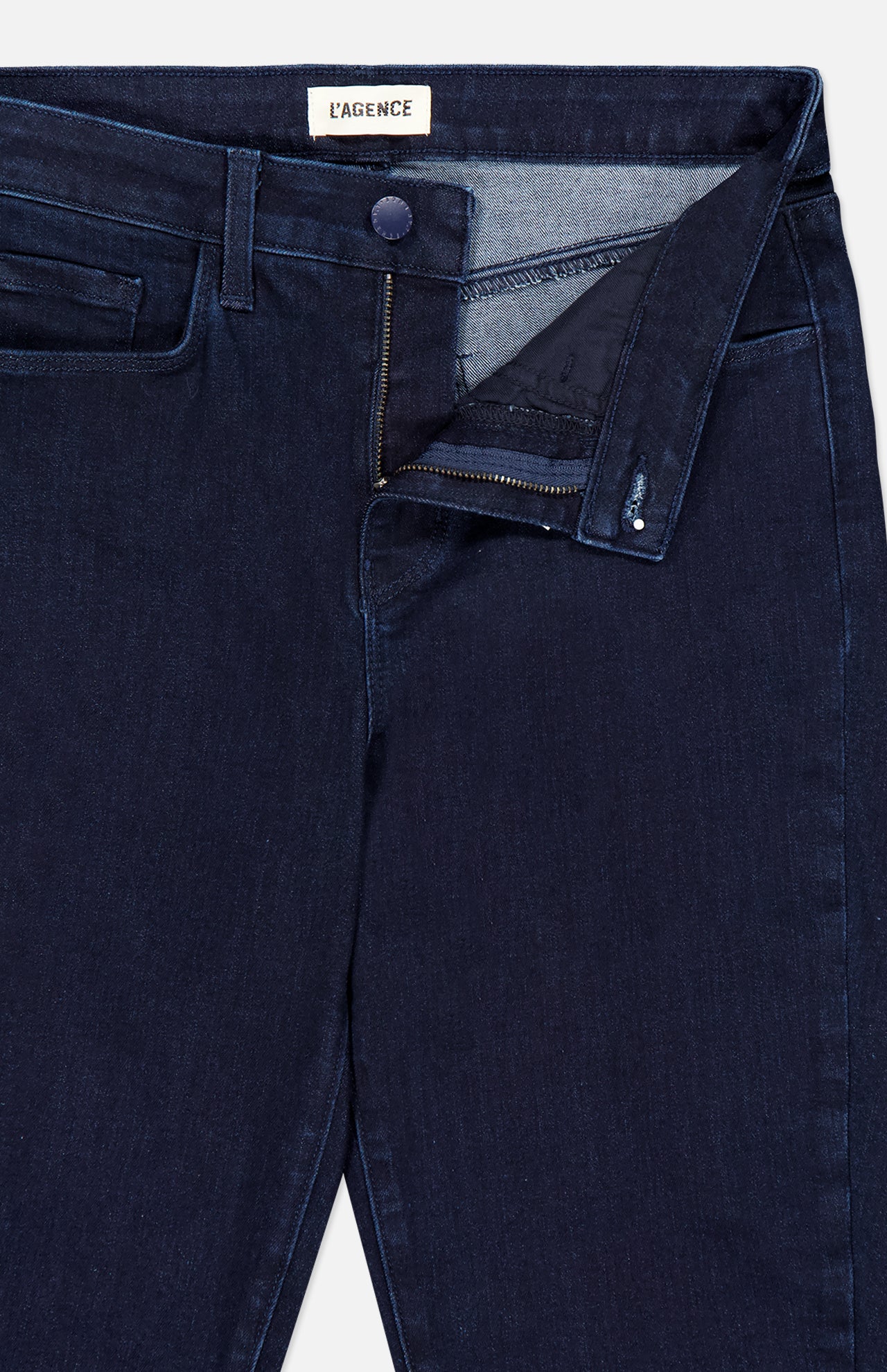 Close-up of Marguerite High Rise Skinny Jean in dark blue stretch denim with pocket, zipper fly, and button. The waistband is partially open, revealing a brand label reading L'Agence inside.