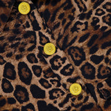 Close-up of the L'AGENCE Nyx Crop Tweed Jacket in brown and black leopard print, detailed with shiny yellow buttons. A partial clothing label appears at the top left.