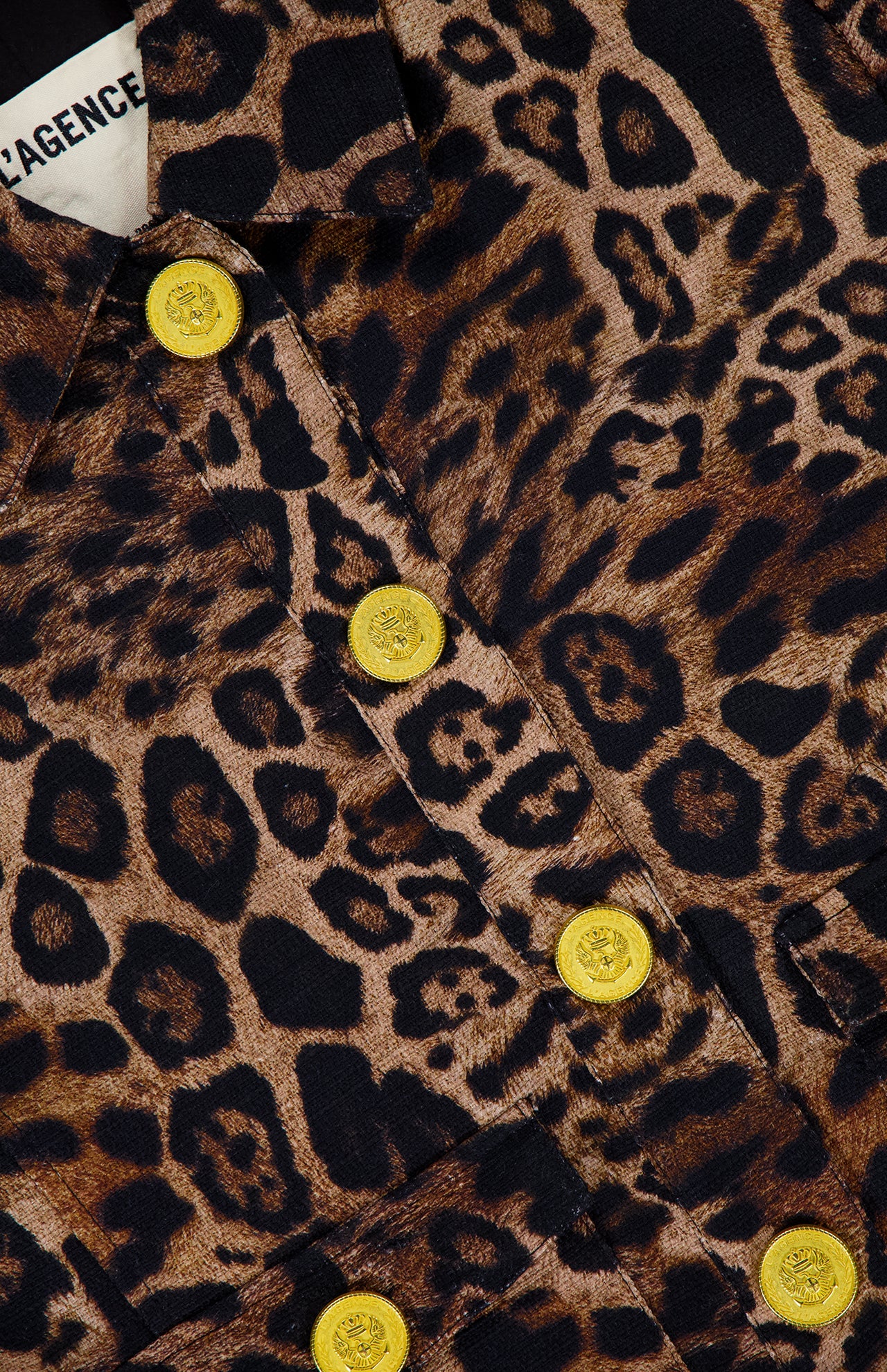 Close-up of the L'AGENCE Nyx Crop Tweed Jacket in brown and black leopard print, detailed with shiny yellow buttons. A partial clothing label appears at the top left.