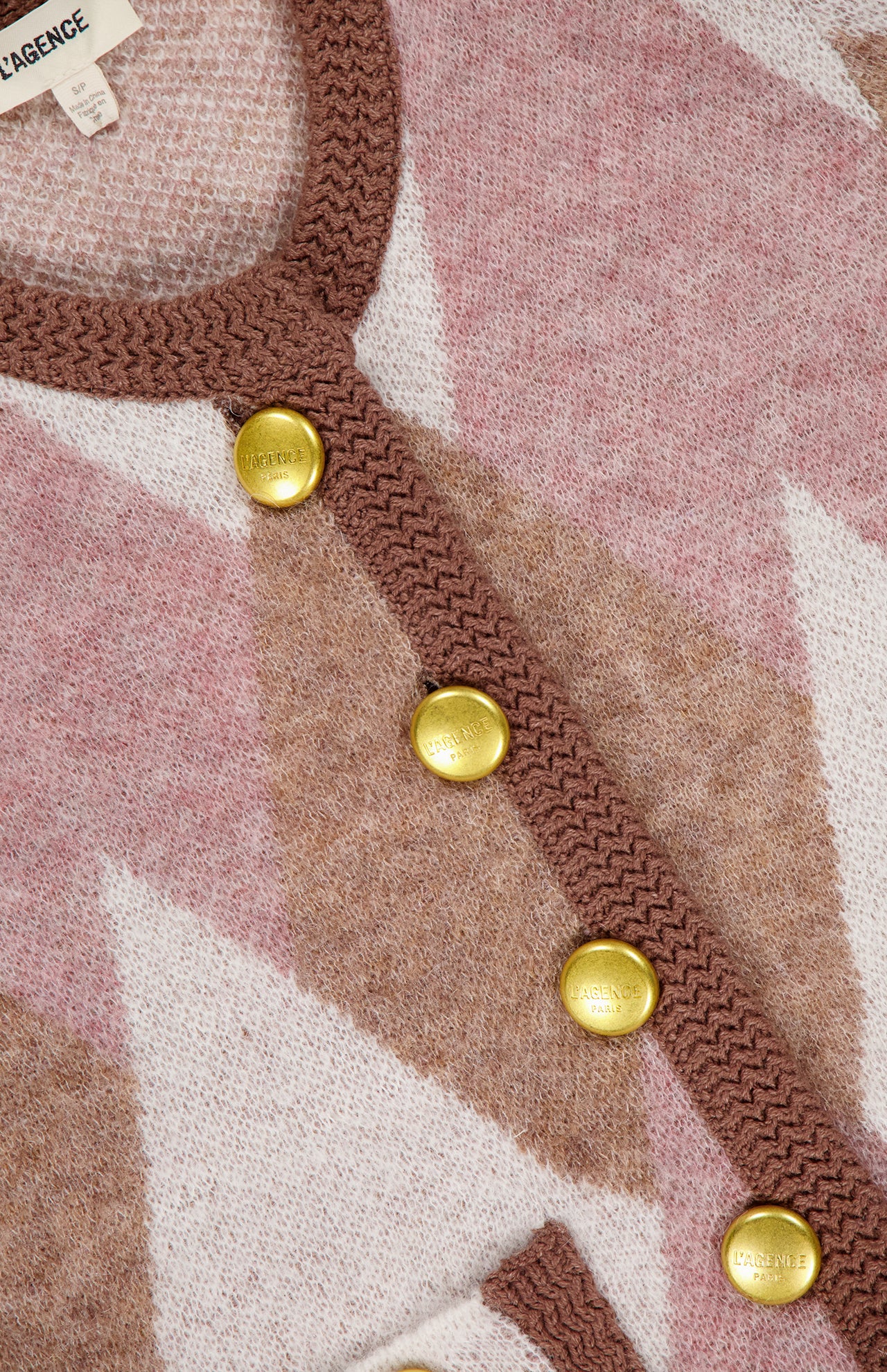 Close-up of the Siobhan Fuzzy Argyle Cardigan by L'AGENCE, featuring a pink and beige geometric pattern, brown trim, and four shiny gold L'AGENCE-engraved buttons. A statement preppy layer with a partially visible label.