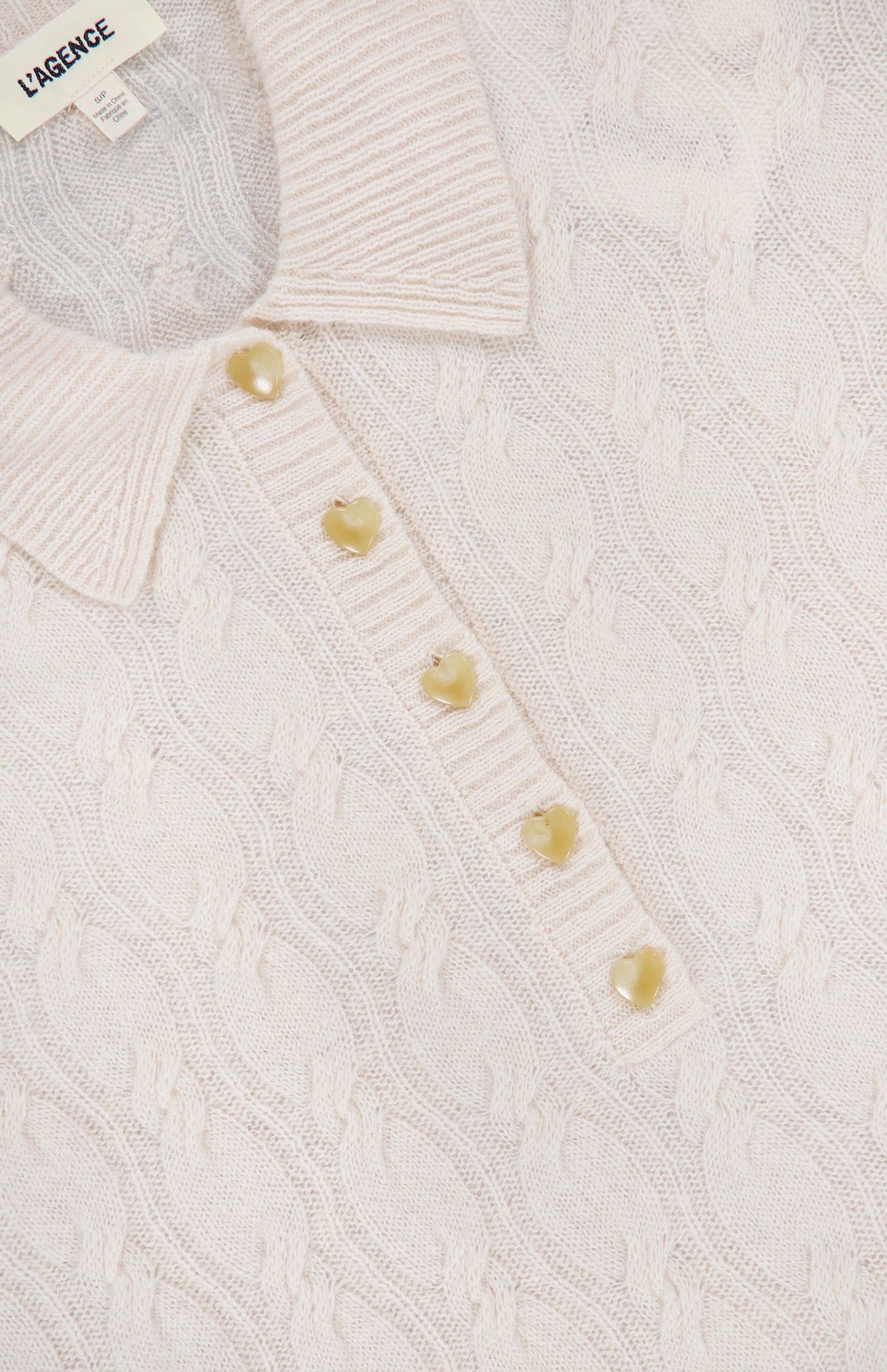 A close-up of the Airy 3/4 Sleeve Heart Button Sweater in a cream ivory hue showcases its ribbed collar and delicate, pale heart-shaped buttons along the placket. The L’AGENCE label is visible inside the collar, adding a timeless element to this elegant sweater.