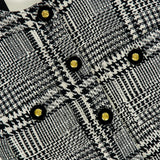 Close-up of the Allegra Short Sleeve Cropped Jacket by L’AGENCE, showcasing a chic black and white plaid textured design with gold and black decorative buttons. The L’AGENCE label is visible, highlighting this sophisticated cropped tweed jacket—an elegant addition to any wardrobe.