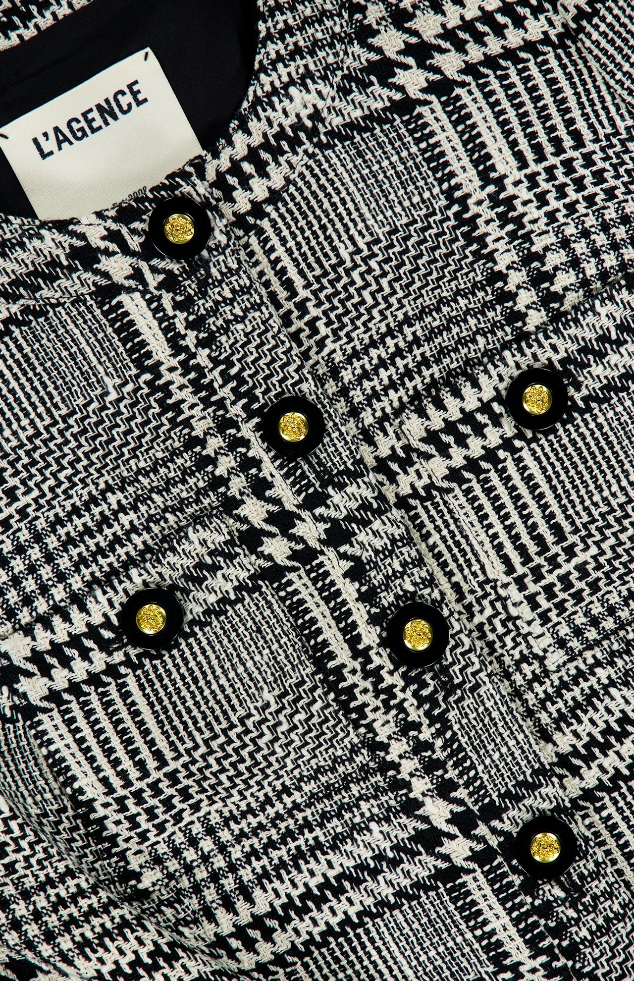 Close-up of the Allegra Short Sleeve Cropped Jacket by L’AGENCE, showcasing a chic black and white plaid textured design with gold and black decorative buttons. The L’AGENCE label is visible, highlighting this sophisticated cropped tweed jacket—an elegant addition to any wardrobe.