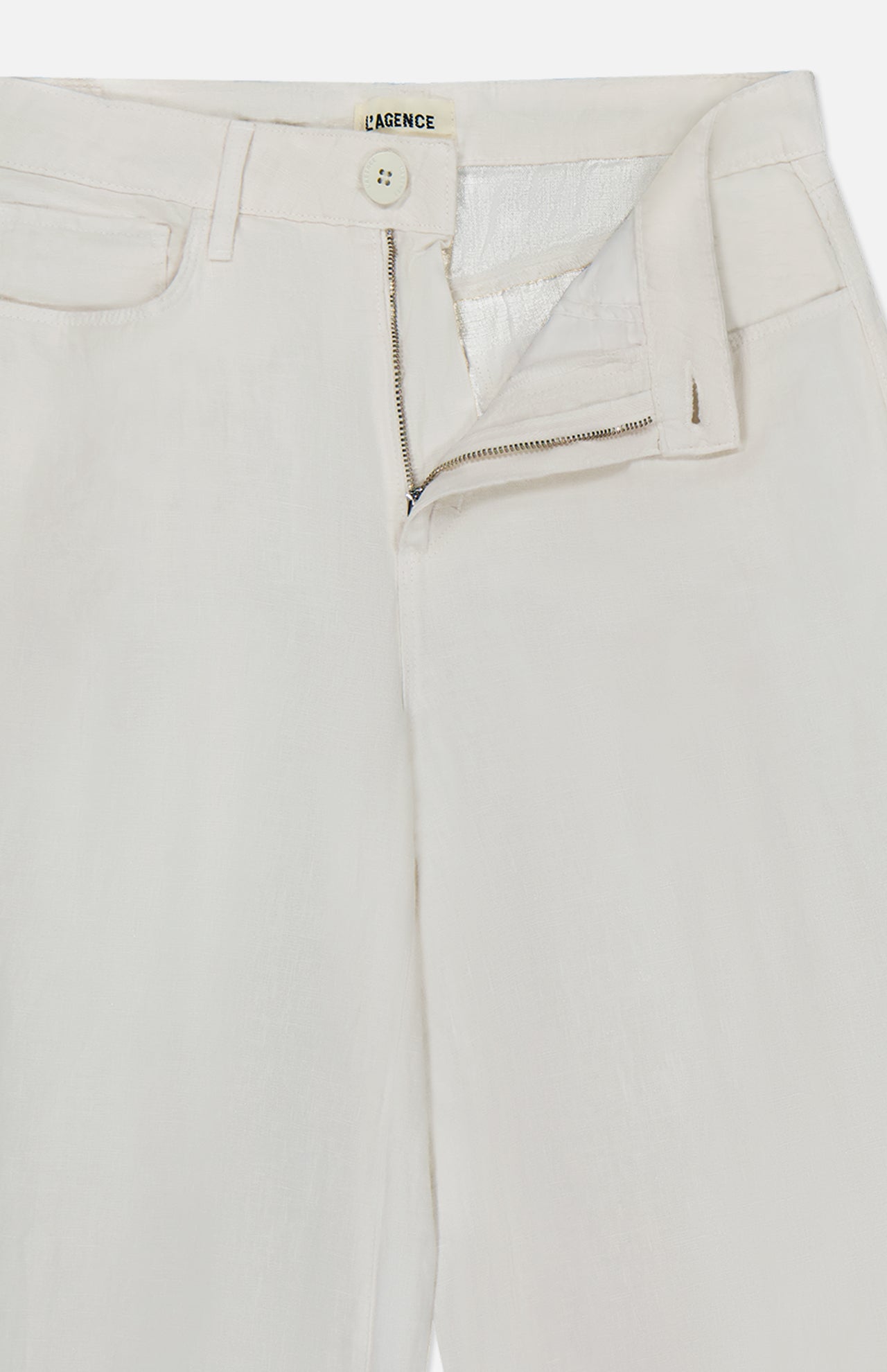 A close-up of the L'Agence Amara High Rise Wide Leg Pant in white linen shows a partially open zipper and button, revealing the waistband and inner tag. The lightweight, textured fabric is tailored for a flattering wide-leg silhouette.