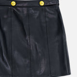 The “Arlena Whipstitch Mini Skirt” by L’AGENCE is a black vegan leather mini skirt featuring two bright yellow-gold buttons at the waistband, shown against a white background.