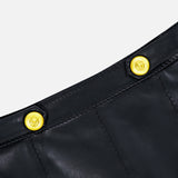 Close-up of the Arlena Whipstitch Mini Skirt by L’AGENCE, crafted from sleek black leather with visible whipstitch detailing and finished with two shiny gold embossed buttons, set against a plain light background.