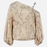 The Asami Chain One Shoulder Blouse by L'AGENCE is a beige, long-sleeve satin top with a chain print, featuring an asymmetrical neckline and loose, gathered cuffs.