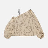 The Asami Chain One Shoulder Blouse by L'AGENCE features a beige chain print, loose asymmetrical sleeve, off-shoulder design, and subtle horsebit accent, displayed on a plain white background.