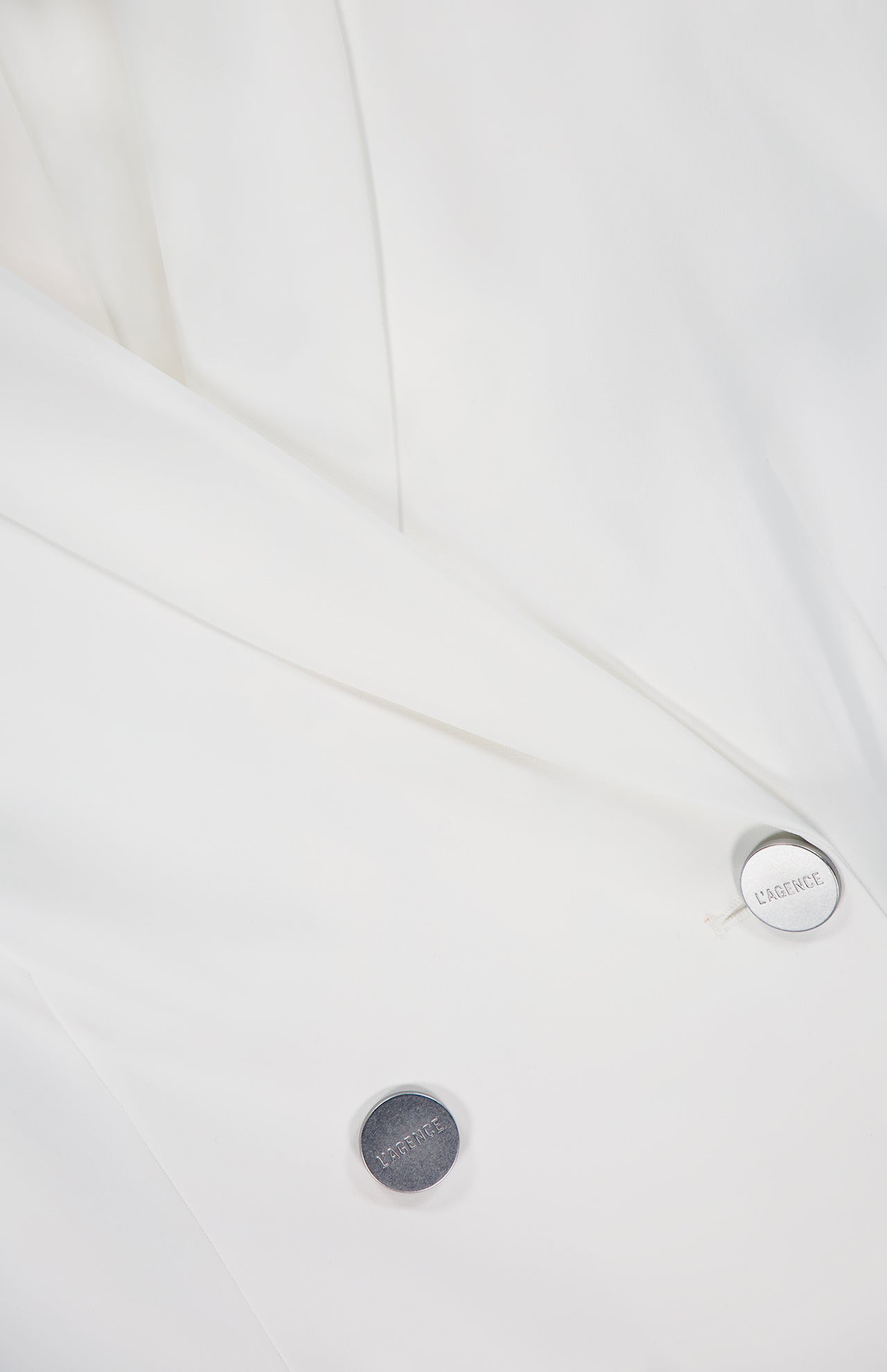 Close-up of the Aven Belted Wrap Blazer by L'AGENCE, showcasing its off-white fabric, smooth texture, clean tailoring, and two metallic buttons—one engraved with ESSENCE.