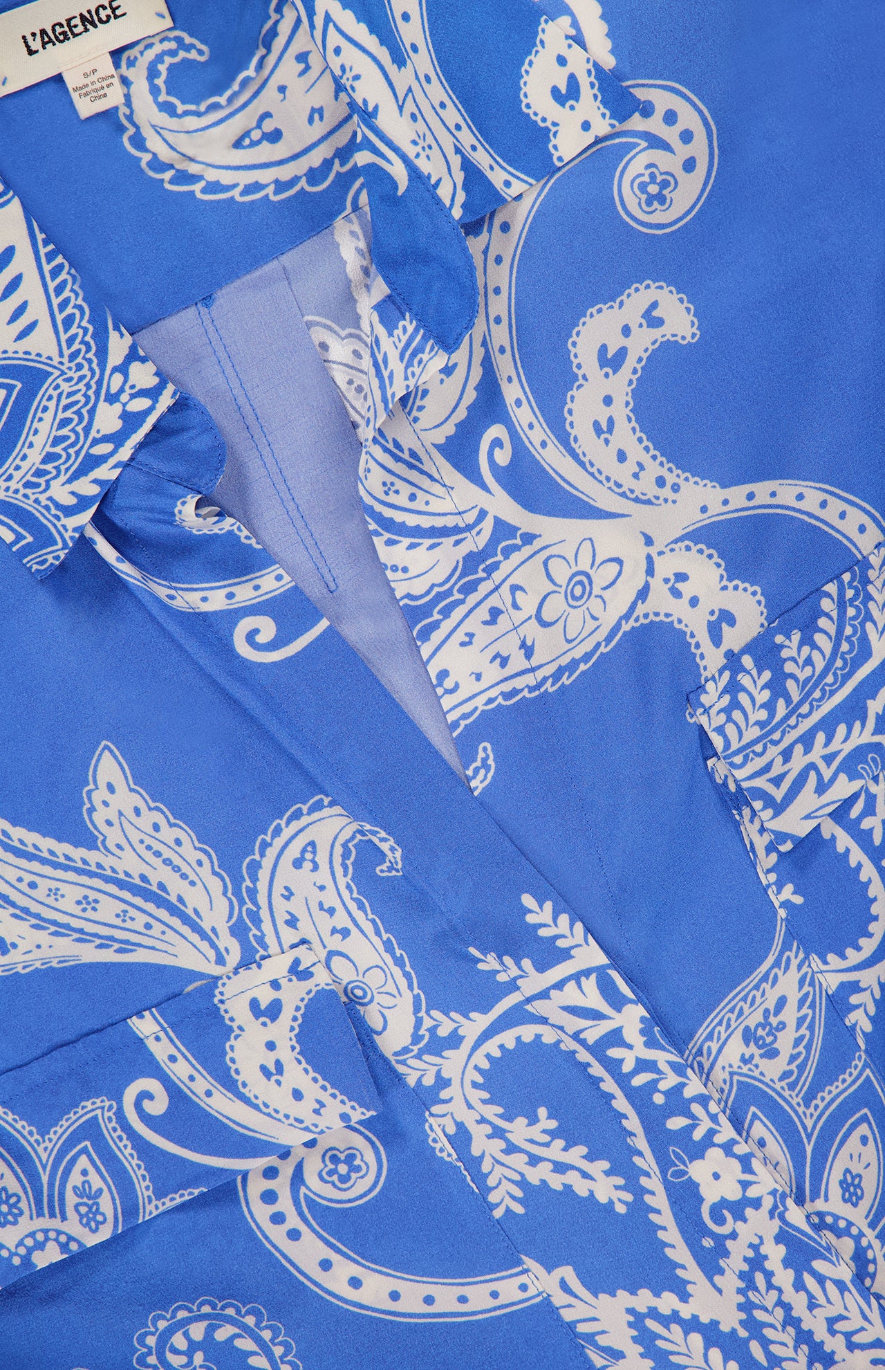 Close-up of the Cameron Long Silk Shirt Dress by L’AGENCE, featuring a blue and white paisley pattern, visible collar, buttons, pocket, and a partially visible brand label in the top left corner.