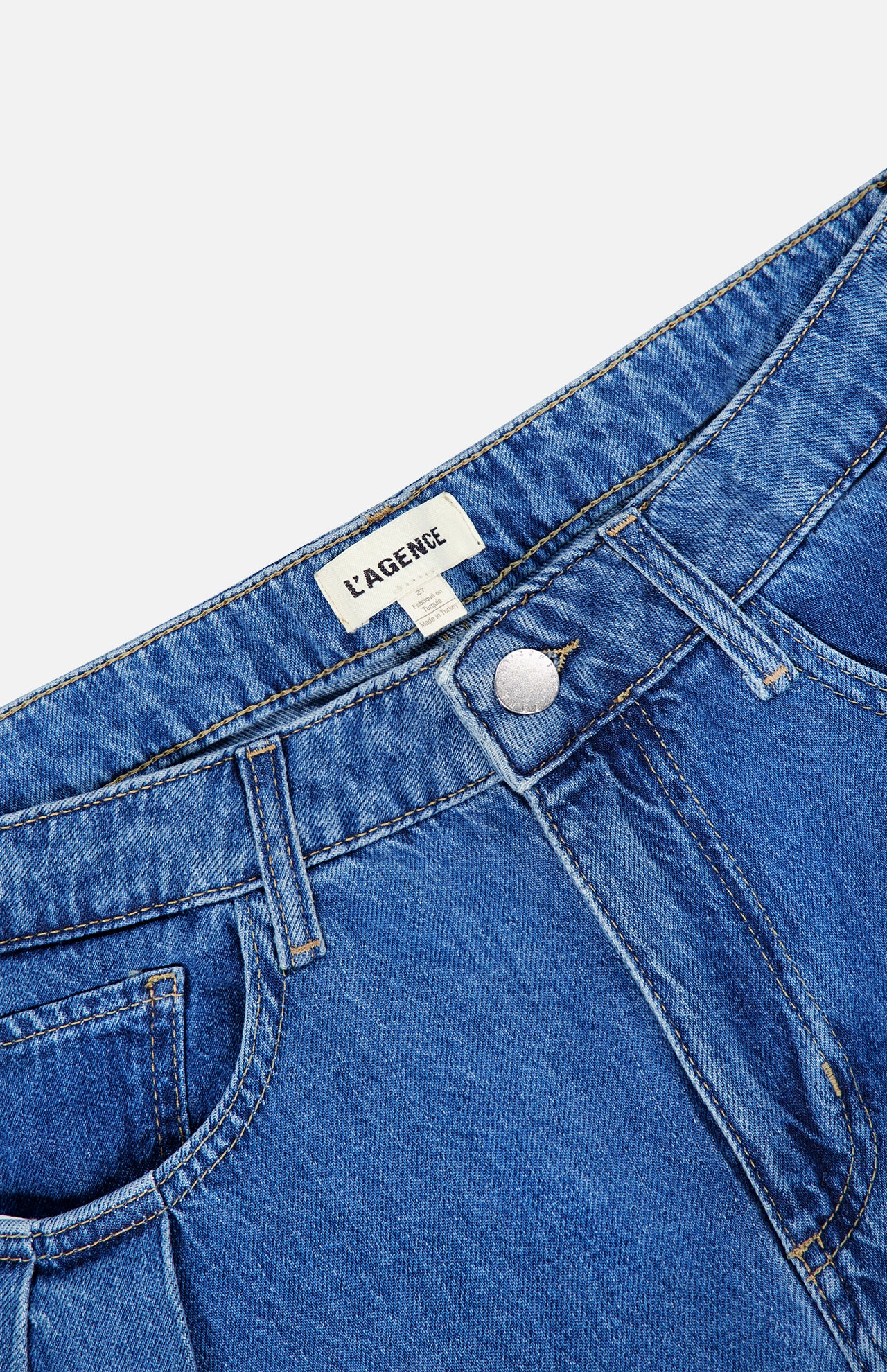 Close-up of the Chayse High Rise Super Wide Leg Jean in Towson, featuring blue denim with a front button closure, belt loops, and visible stitching. A white label with the brand name L’AGENCE is sewn inside the waistband.