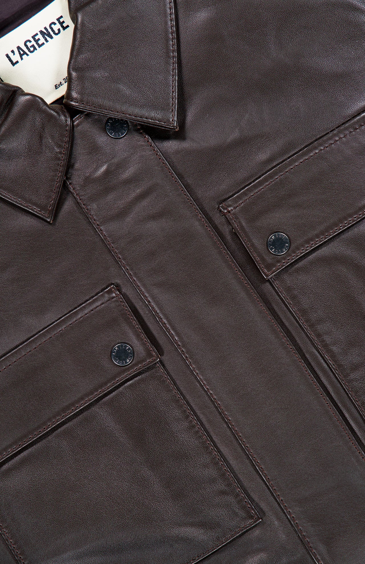 Close-up of the Clotilde Leather Bomber Jacket by L’AGENCE in dark brown, featuring two front flap pockets, black snap buttons, and a visible L’AGENCE label inside the collar.