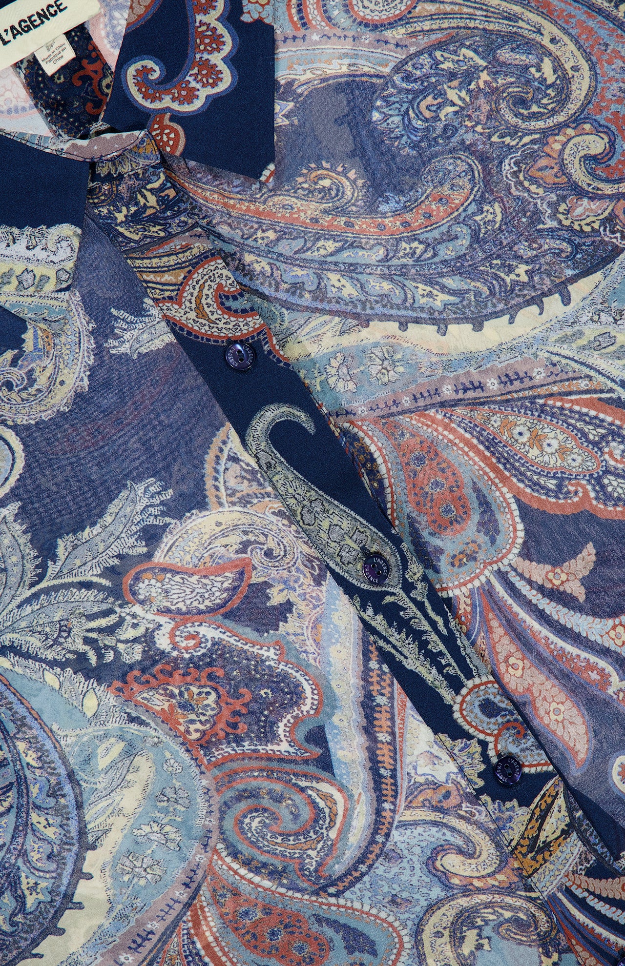 Close-up of the L’AGENCE Cosette Crop Hi Low Shirt, featuring a blue and red paisley pattern, dark buttons, and a pointed collar. The fabric label is visible in the top left corner.