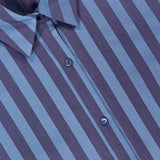 Close-up of the Cosette Cropped Hi Low Shirt by L'AGENCE, featuring vertical dark and light blue stripes, a collar, three visible buttons, and a tag with the L'AGENCE brand name.