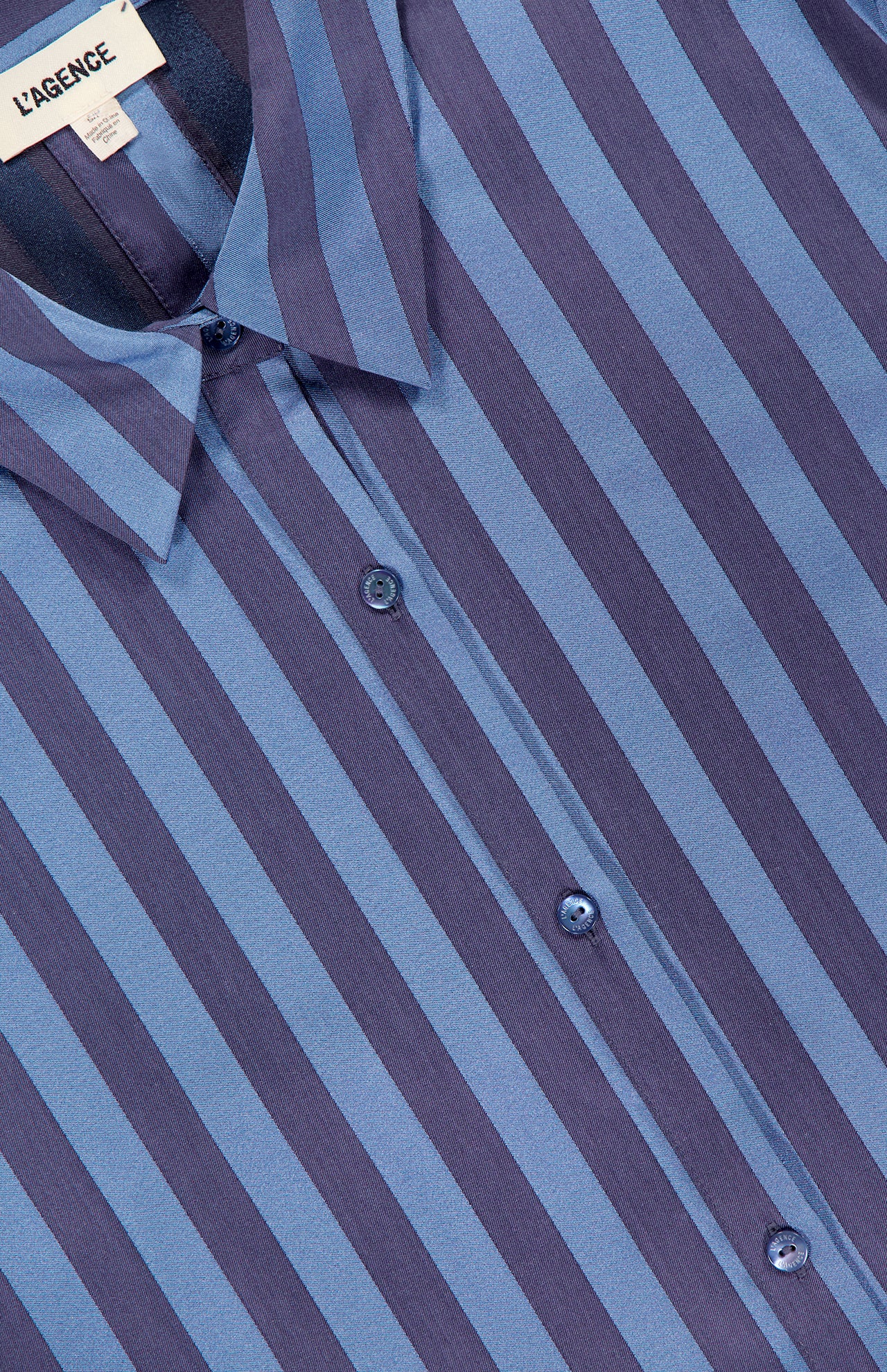 Close-up of the Cosette Cropped Hi Low Shirt by L'AGENCE, featuring vertical dark and light blue stripes, a collar, three visible buttons, and a tag with the L'AGENCE brand name.
