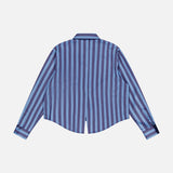The L'AGENCE Cosette Cropped Hi Low Shirt is shown from the back, featuring long sleeves, vertical blue and navy stripes, a collar, buttoned cuffs, and a center-back slit at the hem.