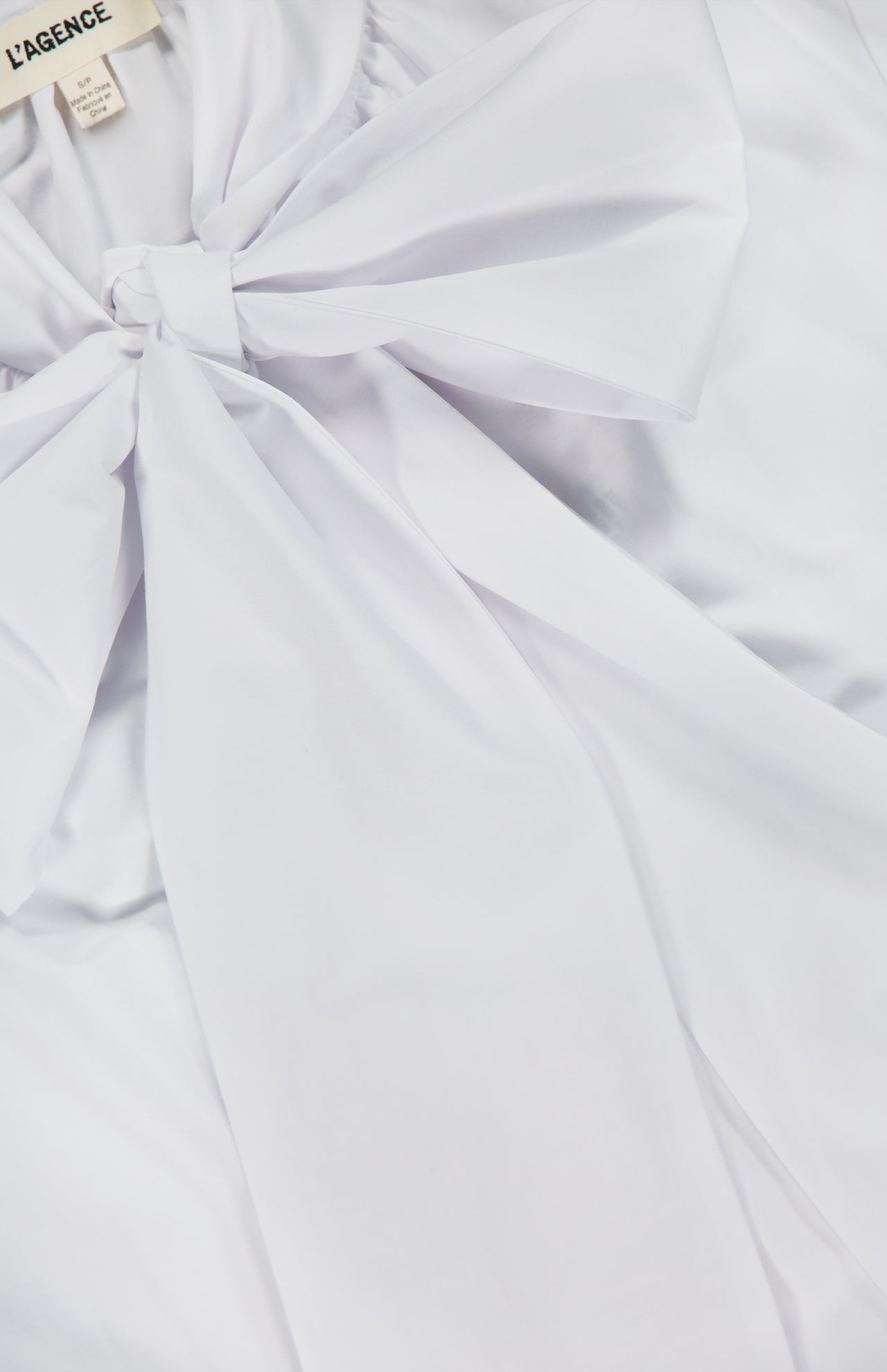 Close-up of the Deon Tie Neck Blouse by L'AGENCE, a white shirt with a large, elegant bow at the collar. The smooth, slightly shiny fabric and signature L’AGENCE tag at the neckline enhance its sophisticated appearance.