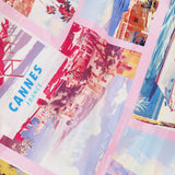 The Driselle Scarf Top by L'AGENCE features a vibrant summer print with vintage Cannes postcard illustrations—palm trees, beaches, and a pool—framed in pink and blue, perfect for your next getaway.