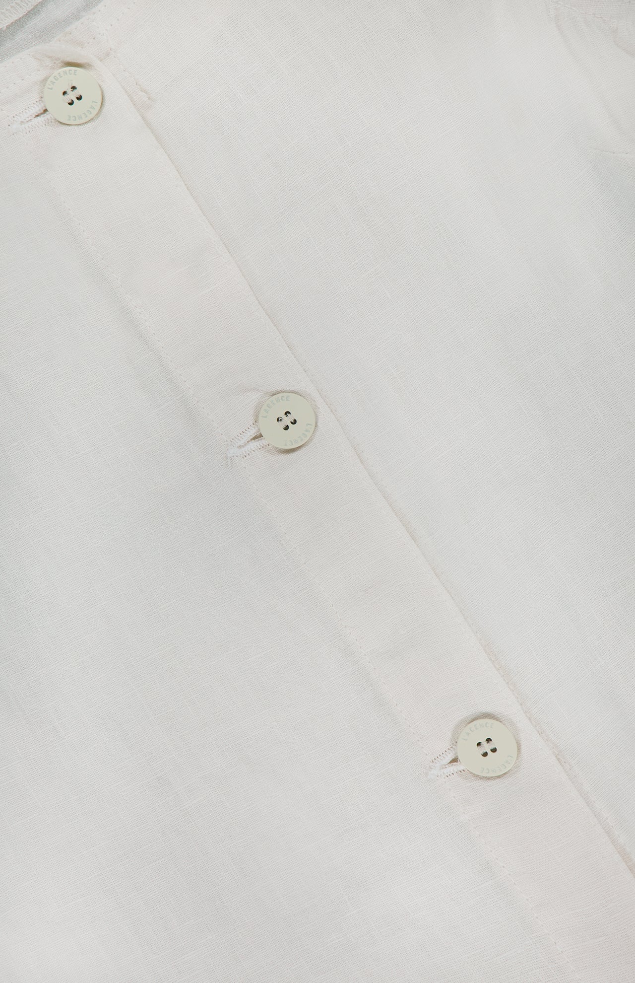 Close-up of the Foster Sleeveless A-Line Top by L'AGENCE in white linen, featuring three white buttons on the front placket with visible fabric texture and stitching details.