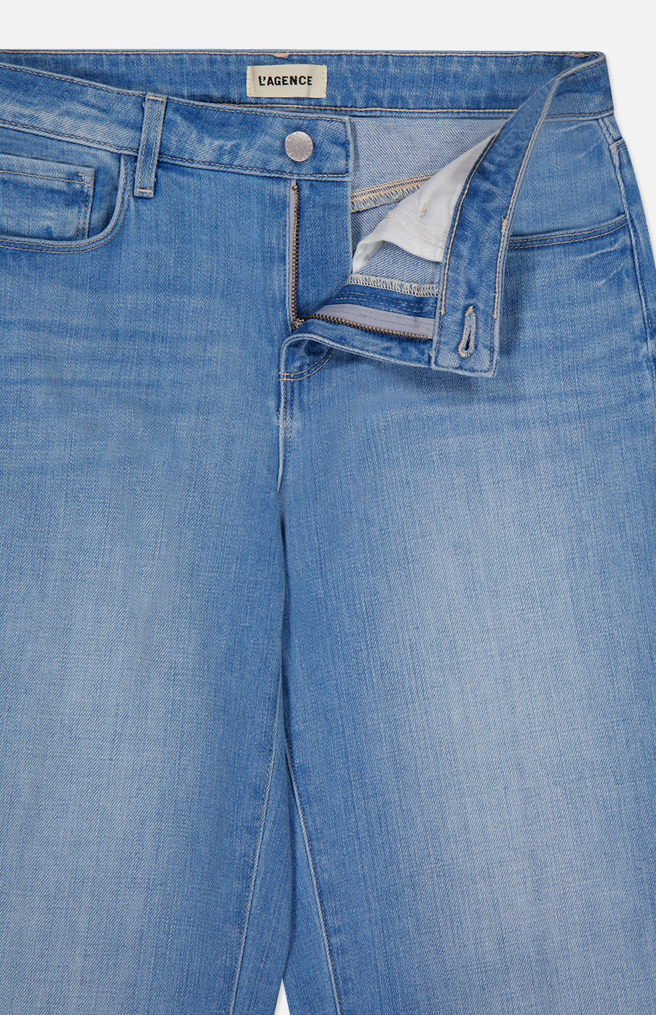 Close-up of L'AGENCE Joanne Low Rise Wide Leg Jean in Pacific, featuring light blue vintage denim with the zipper and button fly open, showing the waistband label and pocket lining.