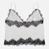 The Kimber Lace Insert Cami by L’AGENCE is a white satin camisole with adjustable spaghetti straps and black lace trim at the neckline, hem, and bust. Photographed on a plain white background.