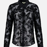 The Laurent Button Up Butterfly Blouse by L’AGENCE is a sheer black statement top with long sleeves, showcasing an all-over butterfly pattern in a striking burnout velvet design and finished with a classic collar, displayed on a plain white background.