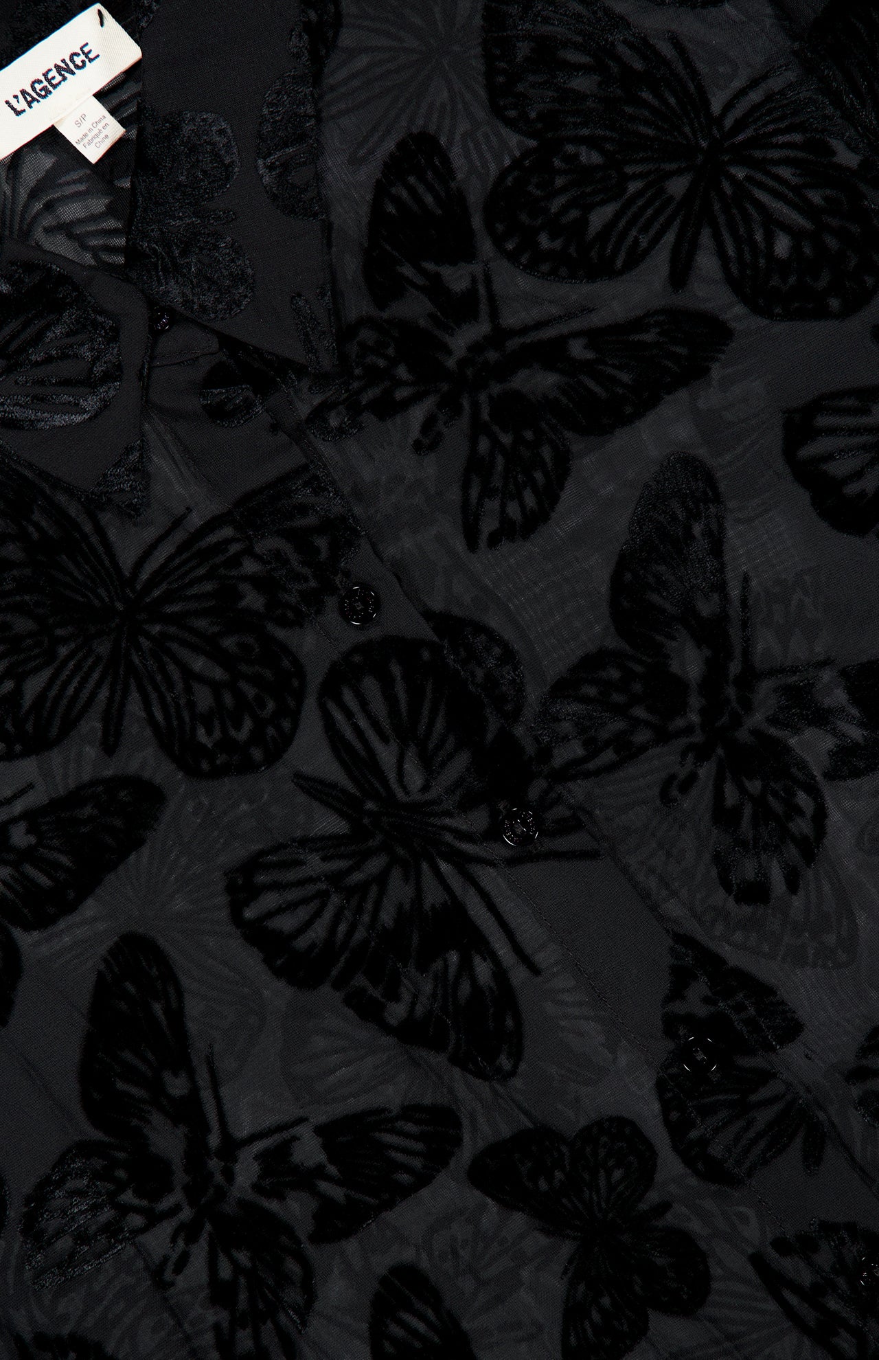 Close-up of the Laurent Button Up Butterfly Blouse by L’AGENCE, featuring sheer black fabric with a velvet butterfly pattern and visible buttons. The top left corner partially reveals the L’AGENCE clothing label, showcasing the exquisite details of this burnout velvet statement blouse.