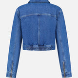 The Micah Oversized Denim Jacket by L’AGENCE is shown from the back in blue denim, featuring long sleeves with zipper details and an elasticized waistband. This jacket boasts visible seam stitching, an oversized fit, and a classic collar for a timeless look.