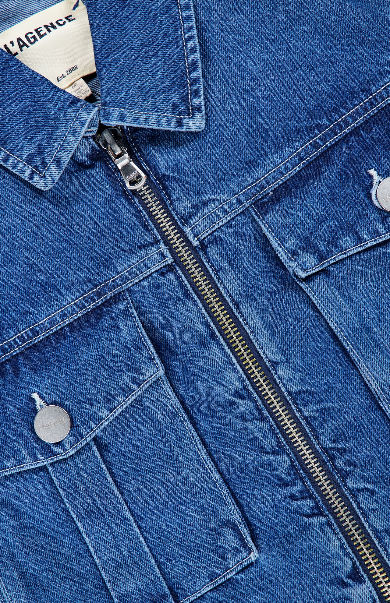 Close-up of the L’AGENCE Micah Oversized Denim Jacket in blue, featuring a front zipper closure and two front pockets with silver buttons. The texture and stitching details of the denim fabric are clearly visible.
