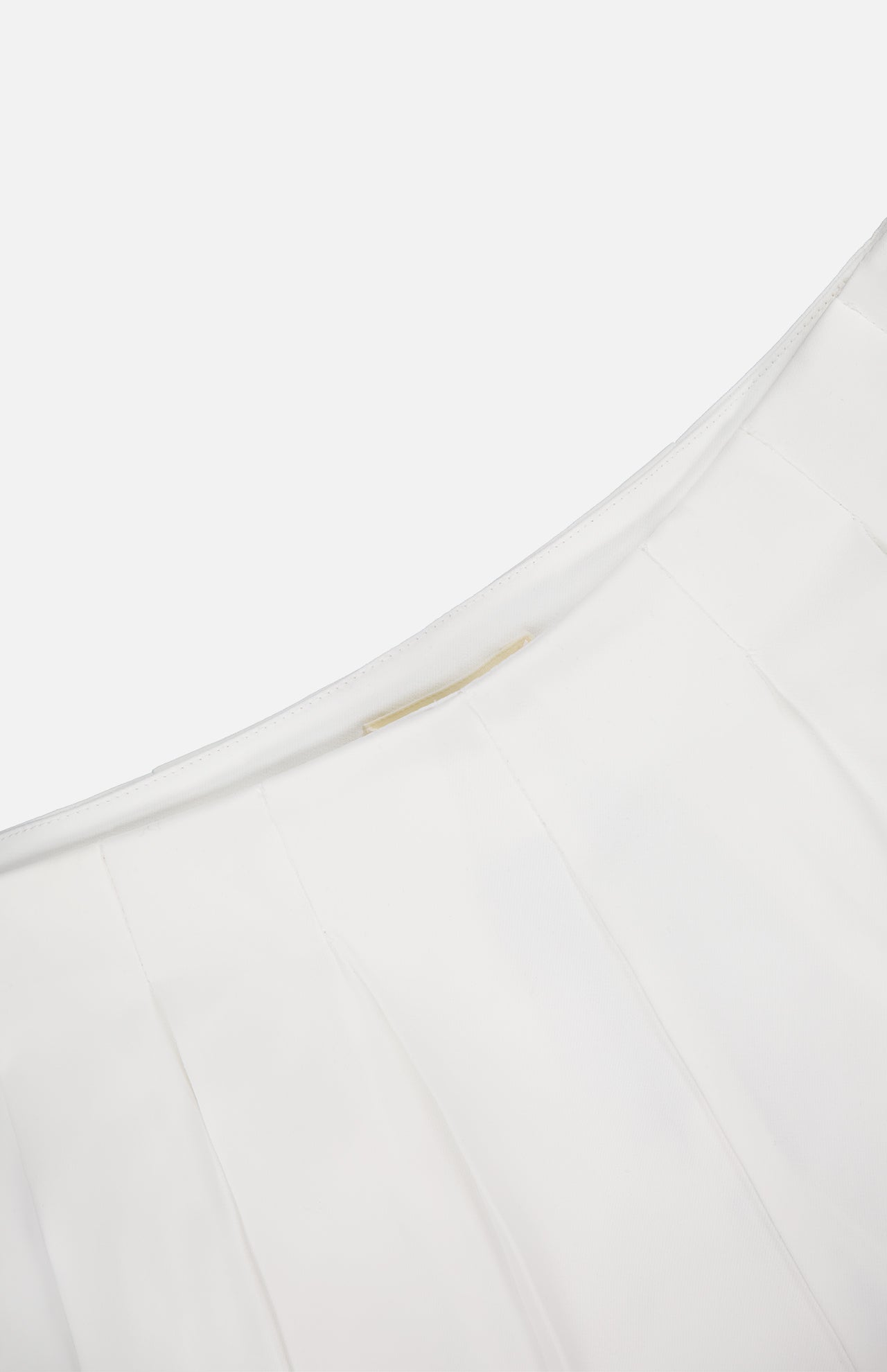 Close-up of the Naamah Pleated Stretch Short by L'AGENCE, featuring wrinkle-resistant pleated white fabric, a curved seam, and a small gold rectangular detail. The plain background highlights this resort-ready essential.