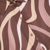 A close-up of the Nina Long Sleeve Swirl Print Blouse by L’AGENCE showcases its elegant geometric wavy pattern in shades of brown, cream, and beige. This button-down top features the L’AGENCE brand label near the collar, highlighting its sophisticated abstract swirl print.