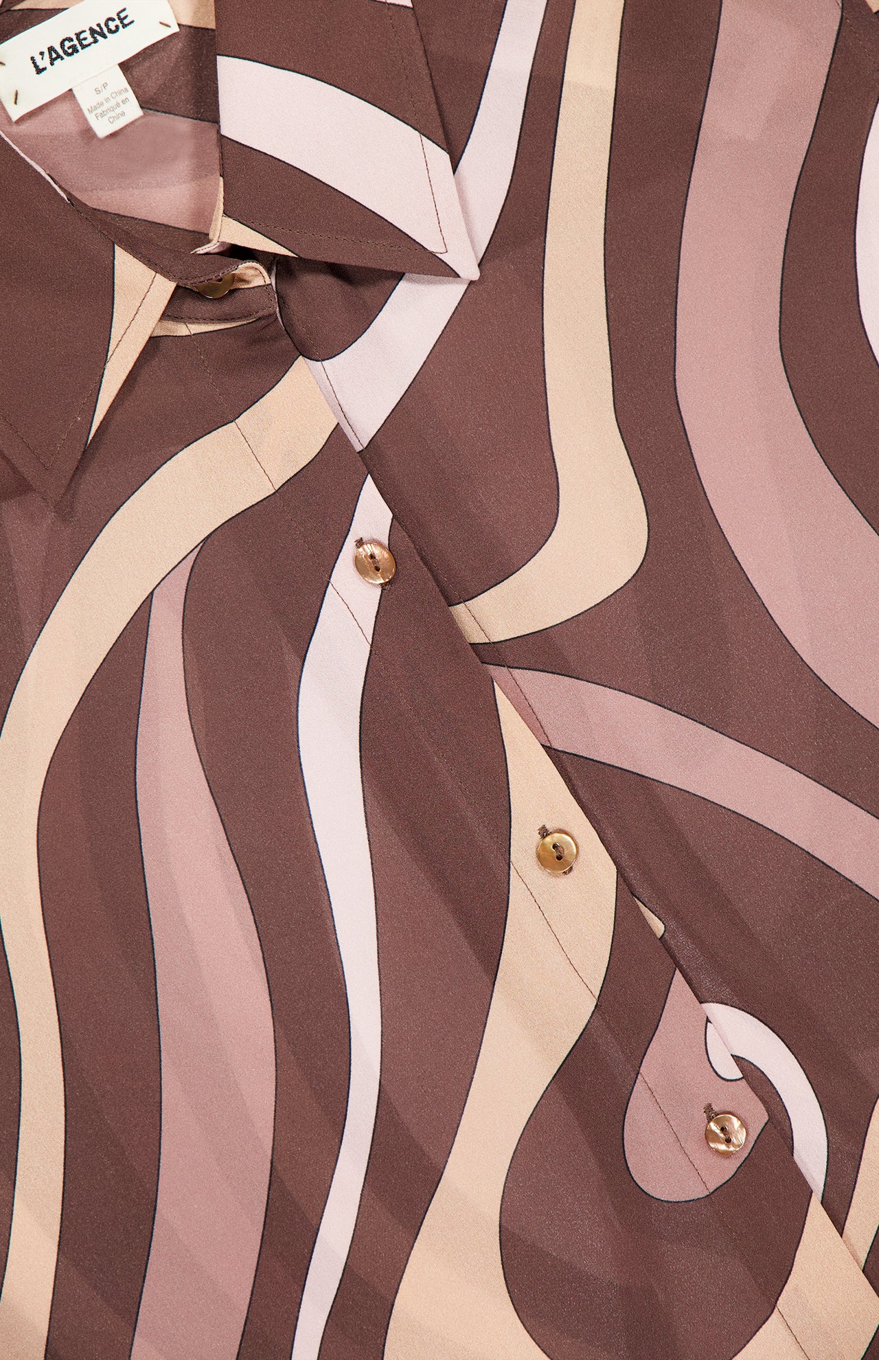 A close-up of the Nina Long Sleeve Swirl Print Blouse by L’AGENCE showcases its elegant geometric wavy pattern in shades of brown, cream, and beige. This button-down top features the L’AGENCE brand label near the collar, highlighting its sophisticated abstract swirl print.