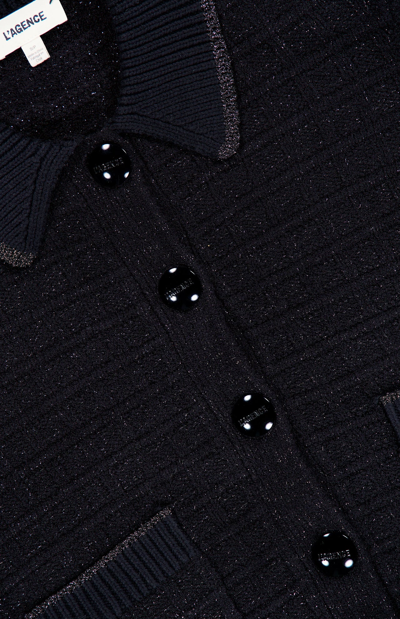 Close-up of the L’AGENCE Novalie Waffle Stitch Short Sleeve Cardigan, showcasing its textured black fabric, four shiny black buttons, and visible pocket—evoking the refined elegance of a classic cardigan. The label near the collar reads L’AGENCE.